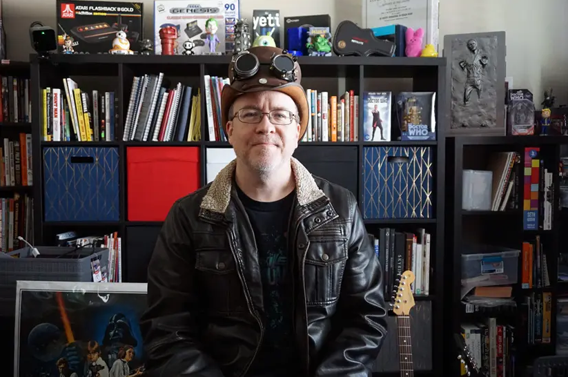 Greg in a steampunk hat, in front of bookshelf of geeky memorabilia